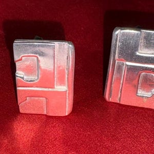May include: Two silver-toned cufflinks with a geometric design. Each cufflink is rectangular with a series of recessed shapes. The cufflinks are displayed on a red fabric surface.