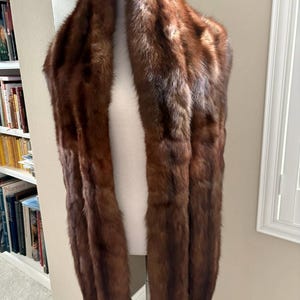 May include: A luxurious brown fur stole displayed on a mannequin. The stole is a rich, warm brown color, with a soft, textured appearance. The fur appears to be in excellent condition, suggesting careful handling and storage. The stole is draped to showcase its length and shape.
