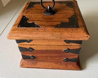 May include: A small, wooden box with a hinged lid and a metal handle. The box is stained a warm brown color and has decorative black metal corner brackets and hinges. The box is rectangular and appears to be handcrafted.