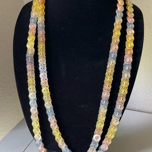 Könnte beinhalten: Eine mehrsträngige Halskette aus pastellfarbenen Knöpfen. Die Knöpfe sind in Gelb-, Rosa- und Blautönen gehalten. Die Halskette wird auf einer schwarzen Büste präsentiert.