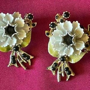 May include: A pair of floral-shaped earrings. Each earring features a white flower with a dark center, surrounded by black beads and gold-colored accents. The earrings have a bow-like design at the bottom and a light green leaf.