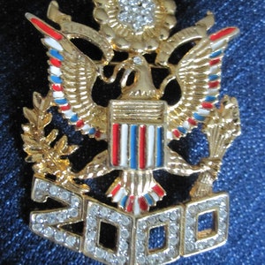 Op de afbeelding: Een gouden broche met een Amerikaanse zeearend met rode, witte en blauwe emaille accenten. De adelaar houdt een bundel pijlen in de ene klauw en een olijftak in de andere. De adelaar zit op een schild met rode en witte strepen. Boven de adelaar bevindt zich een gouden ster met strass steentjes. Onder de adelaar bevindt zich een gouden banner met het jaar "2000" in strass steentjes.