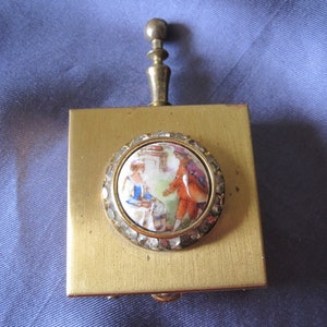 Può includere: Un compatto vintage in ottone con manico decorativo. Il compatto quadrato presenta una scena circolare dipinta a mano di due figure in un giardino, incorniciata da un bordo argentato strutturato. Il compatto è ambientato su uno sfondo blu.