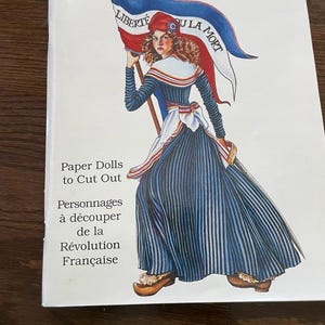 May include: A book titled "The French Revolution" features an illustration of a woman holding a flag with the words "LIBERTE" and "OU LA MORT." The woman is dressed in a blue and white striped dress and a red hat. The book also includes the text "Paper Dolls to Cut Out."