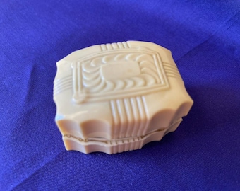May include: A vintage, cream-colored compact with a geometric design on the lid. The compact has a rectangular shape with rounded edges and a decorative pattern. The background is a solid blue fabric.