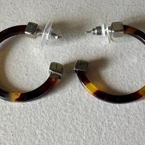 May include: A pair of hoop earrings with a tortoiseshell pattern in shades of brown, yellow, and black. The earrings have a square silver-colored post and a clear plastic backing.