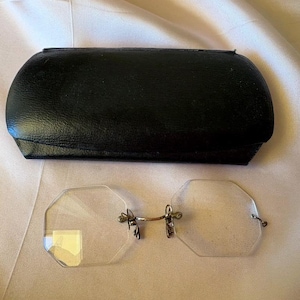 May include: A pair of vintage, rimless eyeglasses with an octagonal lens shape, resting on a light-colored surface next to a black leather eyeglass case. The glasses have a metal bridge and temples.