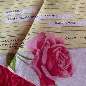May include: A vintage handkerchief with a large pink rose print and green leaves. The handkerchief has text that reads "ARRIVEDERCI", "EUROPA PALACE HOTEL", "ANACAPRI", "MARIO CACACE", and "CAPRI - ITALY". The background is a red fabric.