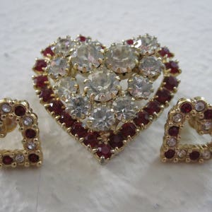 Puede incluir: Un conjunto de joyas en forma de corazón que incluye un broche grande con piedras de imitación rojas y transparentes y dos pendientes más pequeños con piedras de imitación rojas y transparentes.