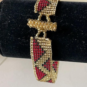 May include: Gold beaded bracelet with red and black detailing. The bracelet has a rectangular design with a central connecting element. The small beads are tightly woven, creating a textured surface. The bracelet is displayed on a black surface.