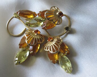 Op de afbeelding: Een broche van goudkleurige metaal met amberkleurige en groene glazen stenen. De broche is gevormd als een tros bloemen met een delicaat, ingewikkeld ontwerp.
