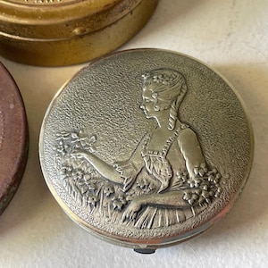 Könnte beinhalten: Eine Sammlung antiker Puderdosen. Die Hauptpuder-Dose ist rund, silberfarben und zeigt ein detailliertes Relief einer Frau in einem historischen Kleid, die Blumen hält. Weitere Puderdosen sind sichtbar, darunter eine goldfarbene und eine braune.