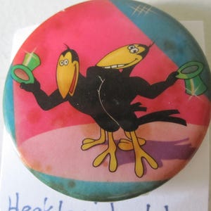 Puede incluir: Un pin redondo con una imagen de dibujos animados de dos pájaros negros con sombreros verdes. Los pájaros están sobre un fondo rosa, azul y verde.