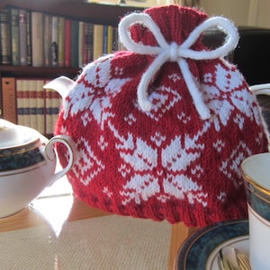 May include: Red and white knitted teapot cozy with a snowflake pattern and white ribbon ties.