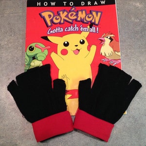 Può includere: Un paio di guanti neri senza dita con polsini rossi. I guanti sono appoggiati su un libro intitolato "How to Draw Pokemon Gotta catch 'em all!" con un'illustrazione di Pikachu giallo.