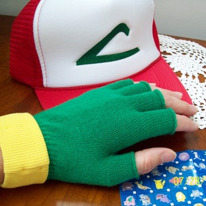Könnte beinhalten: Grüne fingerlose Handschuhe mit gelbem Bündchen, eine rot-weiße Baseballkappe mit grünem 'C'-Logo und ein Pokemon-Sammelkartenblatt.