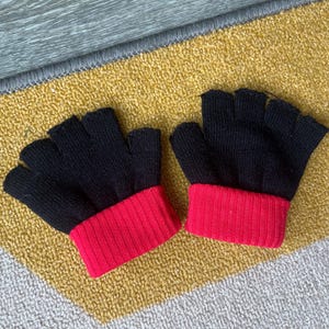 Puede incluir: Un par de guantes sin dedos, negros con puños acanalados rojos. Los guantes están hechos de material de punto y se exhiben sobre una superficie estampada amarilla y gris. Estos guantes están diseñados para mantener las manos calientes mientras dejan los dedos libres.