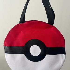 May include: A round, red, white, and black bag with a black handle. The bag is designed to resemble a Pokeball, with a red top, white bottom, and a black stripe and circle in the center. The bag is held by a black handle.