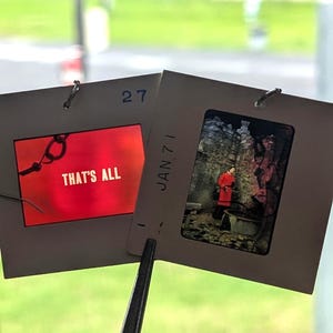 May include: Two vintage slide film frames are held by a metal tool. One frame has the text "THAT'S ALL" on a red background. The other frame shows a person in a red coat. The date "JAN 71" is visible.