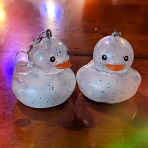 May include: A pair of white glitter rubber ducky earrings with silver hooks. The ducks have blue eyes and orange beaks.