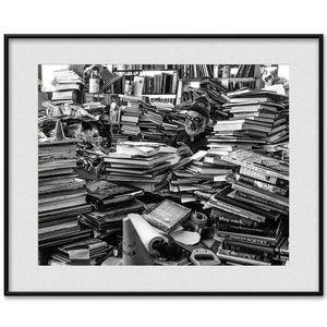 May include: A man sits surrounded by stacks of books in a black and white photo. The books are piled high and reach to the ceiling. The man is wearing a hat and glasses and is looking down at the books. The photo is taken from a slightly elevated angle, giving the viewer a good view of the man and the books.