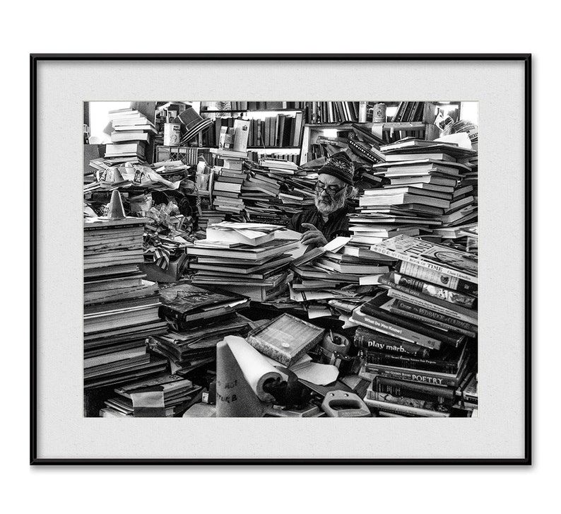 May include: A man sits surrounded by stacks of books in a black and white photo. The books are piled high and reach to the ceiling. The man is wearing a hat and glasses and is looking down at the books. The photo is taken from a slightly elevated angle, giving the viewer a good view of the man and the books.