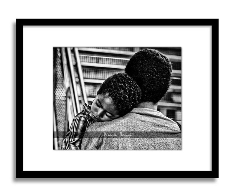 May include: A black and white photograph of a man holding a young child who is sleeping on his shoulder. The child's head is resting on the man's shoulder, and the man's arm is wrapped around the child's back. The man is looking straight ahead, and the child's face is turned to the side. The photo is taken from a low angle, and the background is blurred.