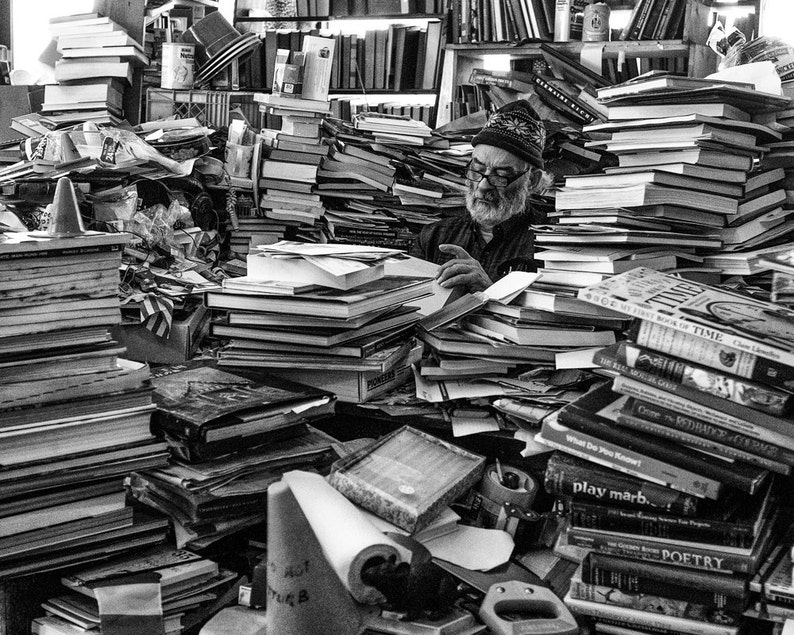 May include: A black and white photo of a man sitting in a room surrounded by stacks of books. The man is wearing a hat and glasses and is looking at the camera. The books are stacked high and are all different sizes and shapes.