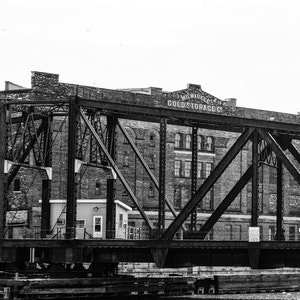 Puede incluir: Una foto en blanco y negro de un puente sobre un río. El puente está hecho de acero y tiene un letrero que dice "Milwaukee Cold Storage Co."