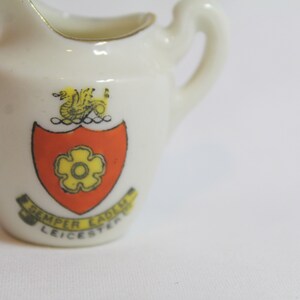 May include: Small, white ceramic pitcher with a handle and a gold rim. The pitcher features a crest with a yellow flower on a red shield, a gold griffin, and the text "Semper Eadem Leicester".