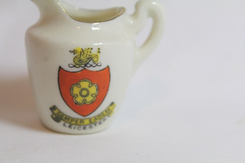 May include: Small, white ceramic pitcher with a handle and a gold rim. The pitcher features a crest with a yellow flower on a red shield, a gold griffin, and the text "Semper Eadem Leicester".