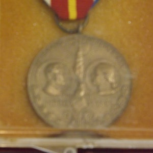 May include: A bronze medal with two profiles of people on either side of a torch. The medal is in a plastic display case with a red, white, and yellow ribbon.