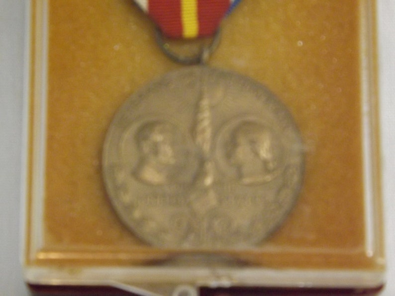 May include: A bronze medal with two profiles of people on either side of a torch. The medal is in a plastic display case with a red, white, and yellow ribbon.