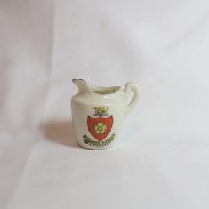 May include: A small, off-white ceramic creamer with a handle and a spout. It features a crest with a red and yellow design and the words "Semper Eadem Leicester". The rim is trimmed with gold.