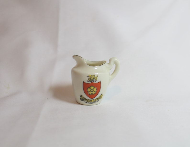 May include: A small, off-white ceramic creamer with a handle and a spout. It features a crest with a red and yellow design and the words "Semper Eadem Leicester". The rim is trimmed with gold.