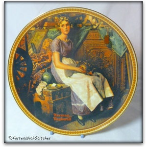 May include: A vintage style collectible plate featuring a painting of a young girl sitting on a wooden chest. The girl is wearing a white dress with a purple bodice and holding a small doll. The plate has a gold rim and the artist's signature "Norman Rockwell" is visible in the bottom right corner.