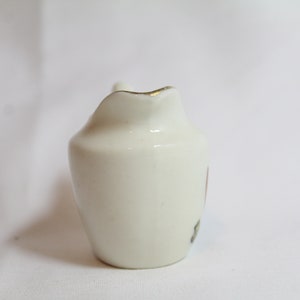 May include: A small, off-white ceramic creamer with a curved spout and a handle. The rim is trimmed with gold. A small floral design is visible on the side. The creamer is likely for serving cream or milk.