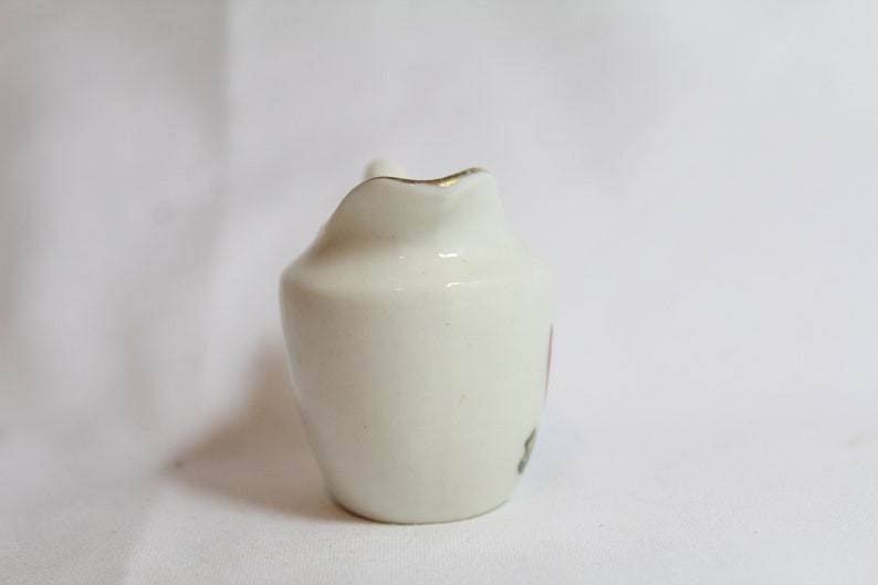 May include: A small, off-white ceramic creamer with a curved spout and a handle. The rim is trimmed with gold. A small floral design is visible on the side. The creamer is likely for serving cream or milk.