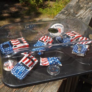 May include: A clear plastic display case containing several miniature toy cars decorated with the American flag design. The cars feature red and white stripes and blue fields of white stars. Also included are two round commemorative coins.