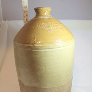 May include: A large, brown stoneware jug with a wide mouth and a rounded body. The jug is glazed in a light brown color and has a darker brown band around the bottom. The jug is labeled "R.W. HOLLAND WINE & SPIRIT MERCHANT MANCHESTER".