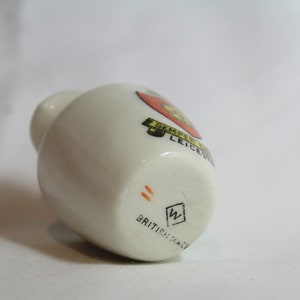 May include: A small, off-white ceramic bottle with a rounded body and a narrow neck. The bottle features a colorful emblem with text and the words "BRITISH MADE" at the base. The emblem includes the text "SEMPER LEICESTER".