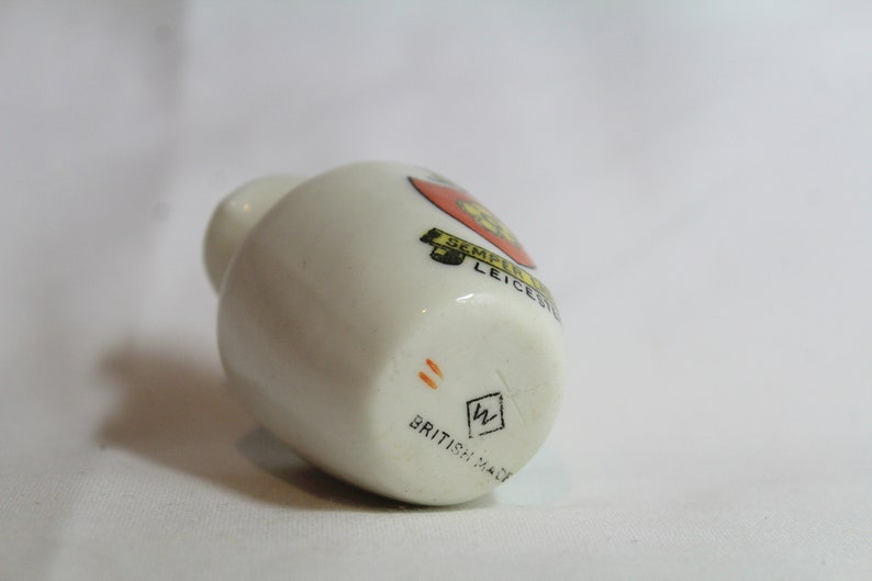 May include: A small, off-white ceramic bottle with a rounded body and a narrow neck. The bottle features a colorful emblem with text and the words "BRITISH MADE" at the base. The emblem includes the text "SEMPER LEICESTER".