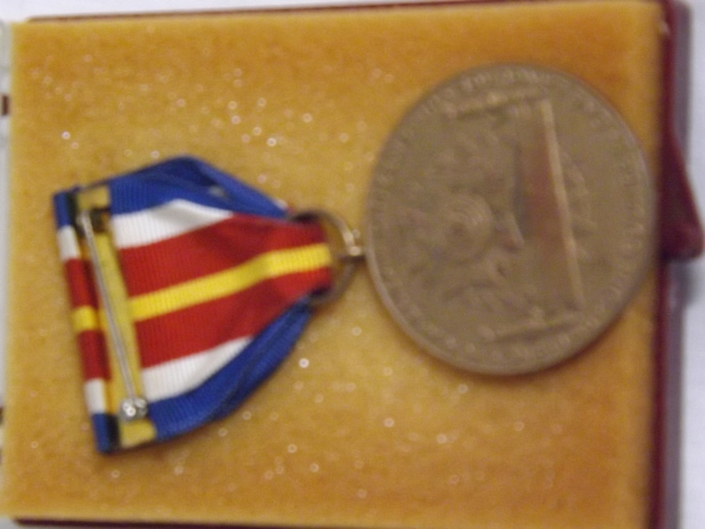 May include: A red, white, and blue ribbon medal with a gold pin and a bronze coin with a design on it. The medal and coin are displayed in a brown velvet box.