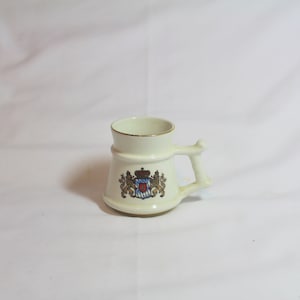 Puede incluir: Una pequeña taza de cerámica blanca con un borde dorado y un diseño de escudo de armas dorado y marrón. La taza tiene un asa.