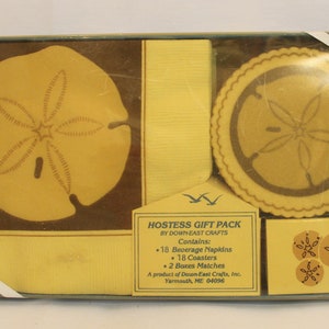 Pode incluir: Um conjunto de presentes para anfitriã da Down-East Crafts. O conjunto inclui guardanapos, porta-copos e caixas de fósforos. Os itens apresentam um design de dólar de areia em tons de amarelo e castanho, embalados numa caixa transparente.