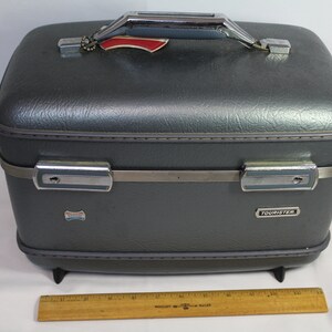 May include: Gray vintage carry-on suitcase with a silver handle and metal accents. The suitcase has the word "Fournister" printed on the front.