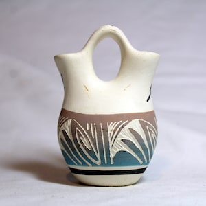 May include: A small, white ceramic pitcher with a double spout and a handle. The pitcher is decorated with a geometric pattern in shades of brown, blue, and white.
