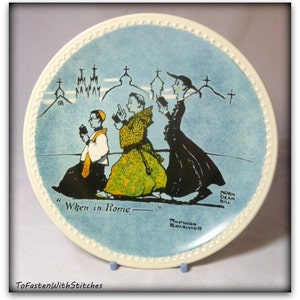 May include: A blue and white ceramic plate with a black and white illustration of three people walking. The plate has a white rim with a beaded design. The text "When in Rome" is printed on the plate. The artist's signature "Norman Rockwell" is printed on the bottom of the plate.
