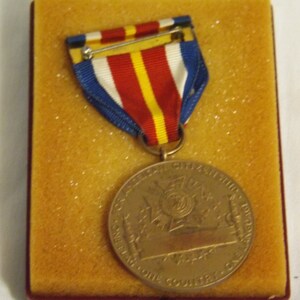 May include: A bronze medal with a red, white, and blue ribbon. The medal features a design of a ship with the text "SERVICE ABOVE SELF" and "ONE NATION ONE COUNTRY ONE CAUSE".