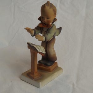 May include: A ceramic figurine of a young boy conducting music. The boy wears a green jacket, brown pants, and a red bow tie. He stands in front of a music stand with sheet music. The figurine is on a rectangular base.